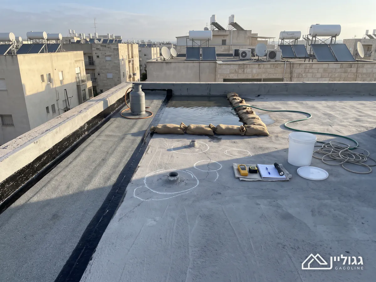 גג שטוח מבטון עם יריעה ביטומנית לאורך המעקה, שק חול סביב שלולית הצפה, צינורות חודרים מסומנים בגיר, מחברת וגלאי לחות על מטלית ודלי לבן.